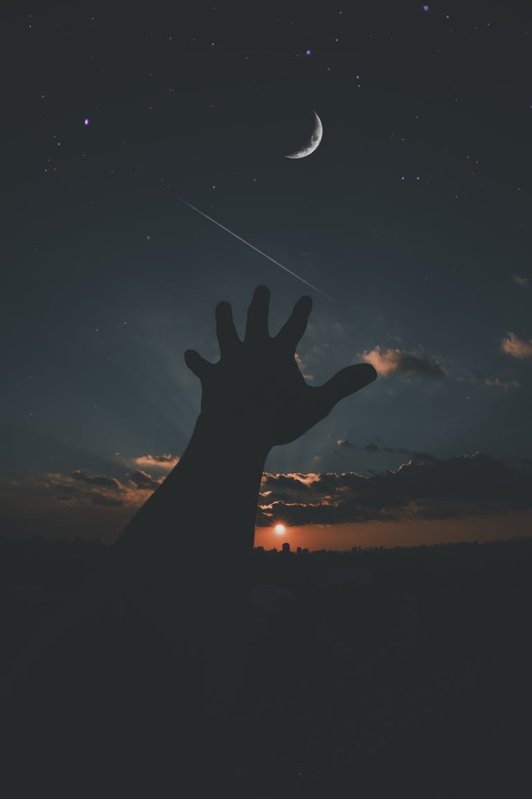 Silhouette of a hand reaching towards the crescent moon and stars in a dramatic dusk sky.